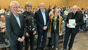 Antonius Kerkhoff, Maria Kröger, Albert Nienhaus, Marianne Heimbach-Steins und Felix Genn stehen nebeneinander in einem holzgetäfelten Raum vor einer Gruppe Menschen, die auf Stühlen sitzen.