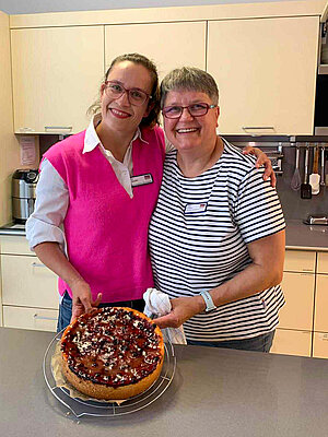 Zwei Frauen haben die Arme umeinander gelegt und halten einen Kuchen in die Kamera, während sie fröhlich lächeln.
