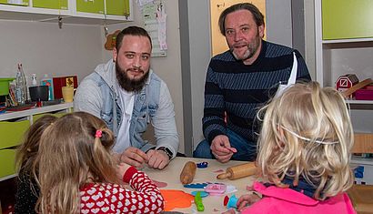 Zwei Männer sitzen an einem kleinen Tisch im Kindergarten, vor ihnen sitzen zwei Kinder mit dem Rücken zum Betrachter.