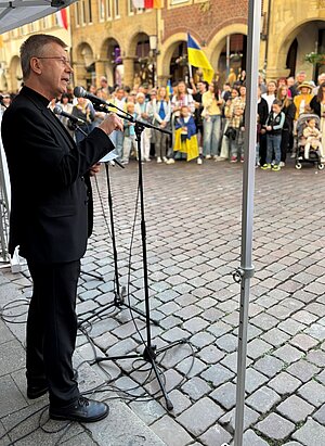 Ein Mann im schwarzen Anzug spricht unter einem Pavillon in ein Mikrofon. Vor ihm steht eine größere Menschenmenge, in der mehrere ukrainische Flaggen zu sehen sind.
