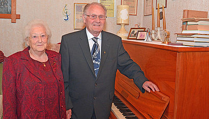 Gertrud und Alfred Giesing stehen in ihrer Wohnung. Er trägt unter dem Anzug eine Krawatte mit Noten und lehnt sich an ein Klavier, an der Wand hängen Urkuden.