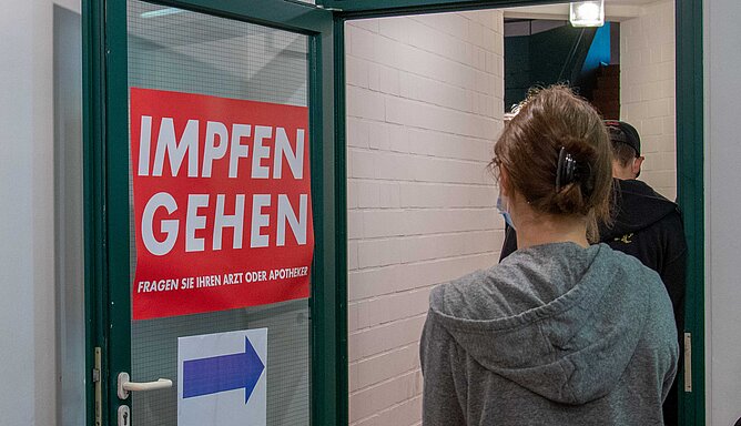 An einer Tür ist das Schild "Impfen gehen" zu lesen, daneben steht eine junge Frau, die nur von hinten zu sehen ist.