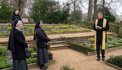Ein Priester steht auf einem Friedhof, links von ihm drei Ordensfrauen.