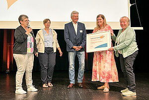 Fünf Personen stehen nebeneinander auf einer Bühne. Die Frau ganz links spricht in das Mikrofon, welches sie in der Hand hält. Die Frau und der Mann in der Mitte lächeln. Der Mann schaut auf den Bilderrahmen, der von den beiden Frauen ganz rechts gehalten wird. Der Bilderrahmen beinhaltet die Urkunde über den gewonnenen Ehrenamtspreis. Fünf Personen stehen nebeneinander auf einer Bühne. Die Frau ganz links spricht in das Mikrofon, welches sie in der Hand hält. Die Frau und der Mann in der Mitte lächeln. Der Mann schaut auf den Bilderrahmen, der von den beiden Frauen ganz rechts gehalten wird. Der Bilderrahmen beinhaltet die Urkunde über den gewonnenen Ehrenamtspreis.