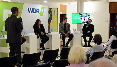 Die Podiumsteilnehmer sitzen vorne nebeneinander.