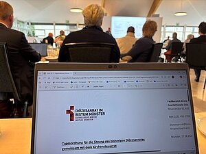 Ein aufgeklappter Laptop steht auf einem Tisch eines Konferenzraums. Auf dem Bildschirm ist die Titelseite einer Präsentation mit dem Logo des Diözesanrats im Bistum Münster und dem Titel „Tagesordnung für die Sitzung des bisherigen Diözesanrates gemeinsam mit dem Kirchensteuerrat“ zu sehen. Im Hintergrund sitzen mehrere Personen an Tischen und blicken auf eine projizierte Präsentation an der Stirnseite des Raums. Der Raum ist hell beleuchtet und verfügt über große Fenster. Ein aufgeklappter Laptop steht auf einem Tisch eines Konferenzraums. Auf dem Bildschirm ist die Titelseite einer Präsentation mit dem Logo des Diözesanrats im Bistum Münster und dem Titel „Tagesordnung für die Sitzung des bisherigen Diözesanrates gemeinsam mit dem Kirchensteuerrat“ zu sehen. Im Hintergrund sitzen mehrere Personen an Tischen und blicken auf eine projizierte Präsentation an der Stirnseite des Raums. Der Raum ist hell beleuchtet und verfügt über große Fenster.