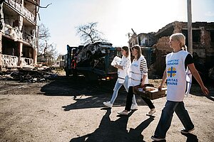 Drei Mitarbeitende von Caritas Ukraine transportieren Lebensmittel in einem kriegszerstörten Wohngebiet. Im Hintergrund sind beschädigte Gebäude, ein Lastwagen und ein ausgebranntes Fahrzeug zu sehen. Drei Mitarbeitende von Caritas Ukraine transportieren Lebensmittel in einem kriegszerstörten Wohngebiet. Im Hintergrund sind beschädigte Gebäude, ein Lastwagen und ein ausgebranntes Fahrzeug zu sehen.