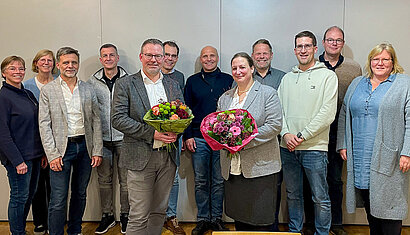 Die beiden gewählten Ehrenamtlichen Timo Brunsmann und Julia Holz stehen mit einem Blumenstrauß in den Händen vor zehn weiteren Personen.