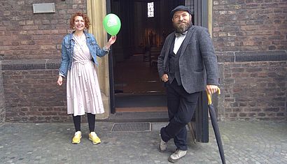 Eine Frau mit einem grünen Ballon in er Hand und ein Mann stehen vor einer offenen Kirchentür.