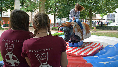 Zwei Mädchen, die mit dem Rücken zur Kamera stehen, beobachten ein anderes Mädchen beim Bullenreiten.