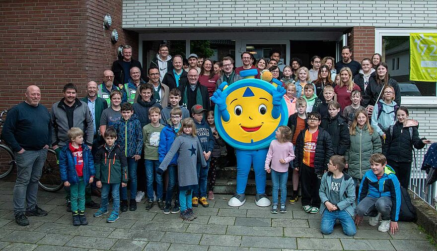 Viele Menschen, insbesondere Kinder, stehen auf einem Gruppenbild neben einem Maskottchen in Form einer Stoppuhr.