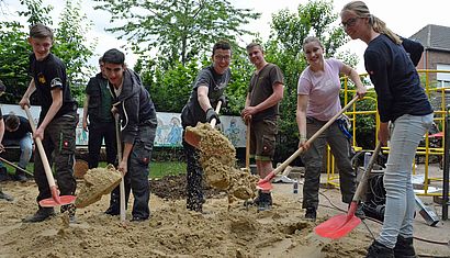 Sechs Jugendliche haben Schaufeln in der Hand, mit denen sie Sand in Richtung des Betrachters schaufeln.