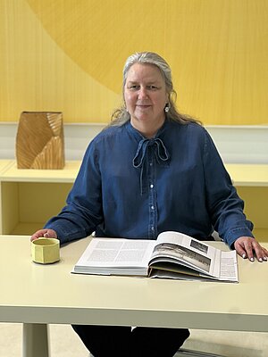 Eine Frau mit langen Haaren sitzt an einem Tisch und blickt ruhig in die Kamera. Sie trägt eine blaue Bluse mit Schleife am Kragen. Vor ihr liegt ein aufgeschlagenes Buch, in der linken Hand hält sie eine helle Tasse. Der Raum ist modern und minimalistisch gestaltet, mit einer gelben Wand im Hintergrund und einem dekorativen Objekt auf einem Regal.