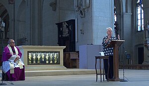 Eine Rednerin steht am Ambo im Altarraum einer großen Kirche. Links von ihr sitzt ein Mann im Messgewand. Eine Rednerin steht am Ambo im Altarraum einer großen Kirche. Links von ihr sitzt ein Mann im Messgewand.