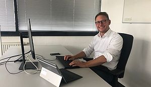 Stephan Dweir sitzt an einem Schreibtisch. Auf dem Tisch befinden sich ein Laptop, daran angeschlossen ein Monitor und eine Tastatur sowie ein Handy.