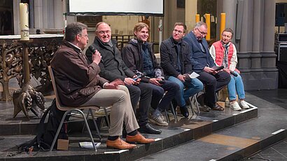 Fünf Männer und eine Frau sitzen vor einem Altar und reden miteinander.