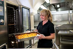 Eva Kemper steht in einer Küche vor einem geöffneten Ofen. Sie hält eine Schale mit einem Auflauf in den Händen.