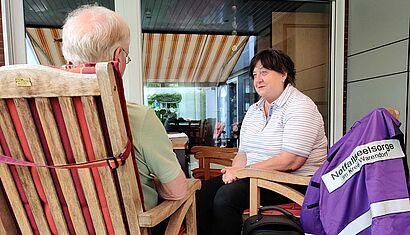 Zwei Personen sitzen auf einer Terrasse, über einer Stuhllehne hängt eine Jacke mit der Aufschrift Notfallseelsorge.