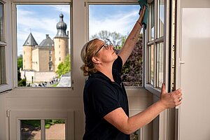 Rieke Schneiders-Verhoolen reinigt einen geöffneten  Fensterflügel. Durch das Fenster ist die Jugendburg Gemen zu sehen..