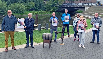 Mehrere Menschen stehen im Freien und halten Plakate hoch, vor ihnen stehen ein Weinfass und ein Holzkreuz, im Hintergrund ist ein Fluss zu sehen.