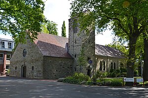 Steingebäude mit Kirchturm und Satteldach, umgeben von großen Bäumen und Grünflächen. Es handelt sich um eine kleine Kirche mit rundbogigen Fenstern und Eingangstüren. Links neben der Kirche ist ein modernes Gebäude mit grauem Dach und roten Backsteinwänden zu sehen. Es ist ein sonniger Tag mit leicht bewölktem Himmel.