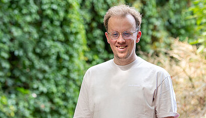 Jakob Rauschel schaut lächelnd in die Kamera und steht vor einer Hecke.