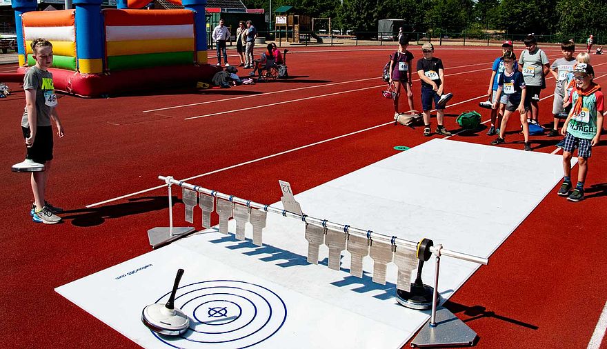 Kinder stehen auf einem Sportplatz und spielen auf einer Kunststoff-Bahn Eisstock-Schießen.