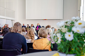 Vor einer Leinwand stehen mehrere Menschen. Eine Frau hat ein Mikrofon in der Hand und spricht zu dem vor ihr sitzenden Publikum.