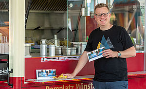 Christoph Aperdannier steht an einem Imbisswagen auf dem Domplatz Münster. In der Hand hält er den Flyer "Gestärkt.Leben".