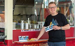 Christoph Aperdannier steht an einem Imbisswagen auf dem Domplatz Münster. In der Hand hält er den Flyer "Gestärkt.Leben". Christoph Aperdannier steht an einem Imbisswagen auf dem Domplatz Münster. In der Hand hält er den Flyer "Gestärkt.Leben".