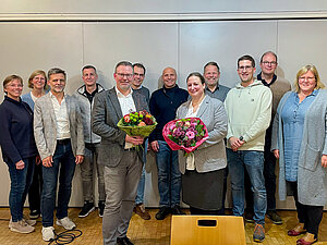 Die beiden gewählten Ehrenamtlichen Timo Brunsmann und Julia Holz stehen mit einem Blumenstrauß in den Händen vor zehn weiteren Personen. 