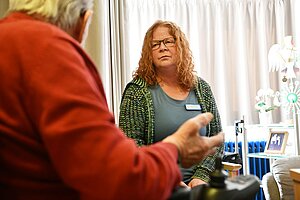 Eine sitzende Person in einem Strickoberteil mit Namensschild befindet sich in einem hellen Raum mit Vorhängen im Hintergrund. Im Vordergrund ist der teilweise sichtbare Oberkörper einer weiteren Person zu sehen, die eine Hand ausgestreckt hält. Rechts im Raum stehen ein Rollstuhl, ein Tisch mit gerahmten Fotos und dekorativen Gegenständen.