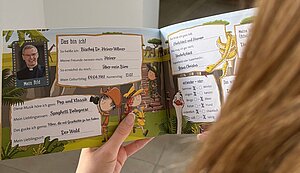 Eine Person hält ein aufgeschlagenes, farbig illustriertes Kinderbuch in den Händen. Auf der Doppelseite ist ein Steckbrief von Bischof Dr. Heiner Wilmer mit Foto und persönlichen Angaben wie Lieblingsmusik, Lieblingsessen und Lieblingsort zu sehen. Eine Person hält ein aufgeschlagenes, farbig illustriertes Kinderbuch in den Händen. Auf der Doppelseite ist ein Steckbrief von Bischof Dr. Heiner Wilmer mit Foto und persönlichen Angaben wie Lieblingsmusik, Lieblingsessen und Lieblingsort zu sehen.