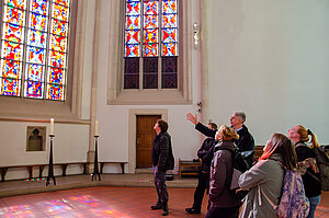 Eine Gruppe von Menschen steht vor bunten Kirchenfenstern und betrachtet sie.
