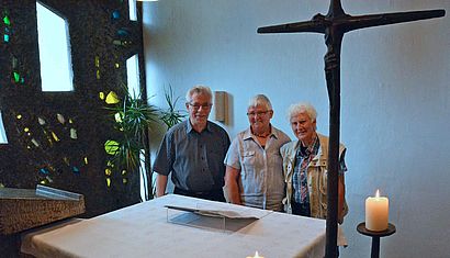 In einer Kapelle stehen hinter dem Altar (von links) Pfarrer Heinrich Bösing, Karin Jäschke und Ursula Weinbrenner. Rechts im Vordergrund steht ein Kreuz, vor dem eine Kerze brennt.