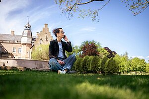 Julia Franzmann sitzt im Schneidersitz auf einer grünen Wiese vor der Jugendburg und telefoniert mit einem Mobiltelefon.