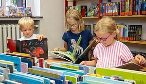 Drei Kinder stehen in einem Raum mit Regale und Kisten, in denen sich Bücher, befinden und lesen.