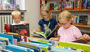 Drei Kinder stehen in einem Raum mit Regale und Kisten, in denen sich Bücher, befinden und lesen. Drei Kinder stehen in einem Raum mit Regale und Kisten, in denen sich Bücher, befinden und lesen.