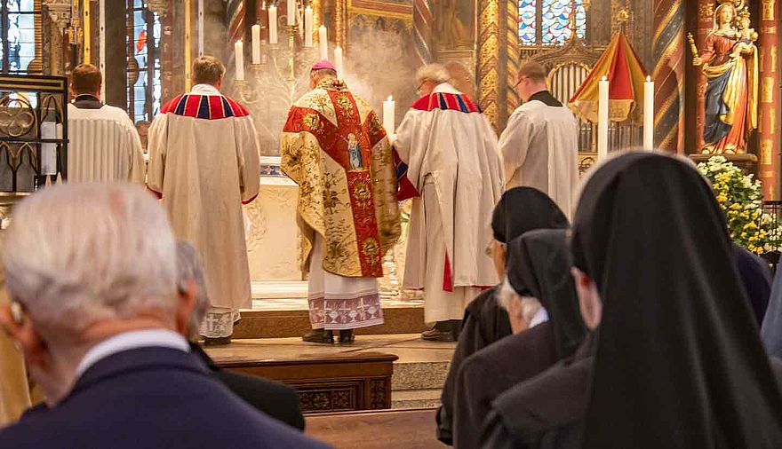 Momentaufnahme aus dem Gottesdienst, im Hintergrund stehen Geistliche am Altar, im Vordergrund sind, mit dem Rücken zur Kamera, Ordensfrauen und ein Mann zu erkennen.