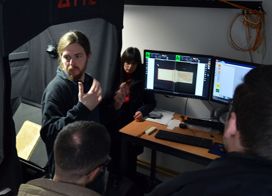 Mehrere Studierende schauen auf zwei Computerbildschirme, auf einem der Bildschirme ist die Abbildung eines Buches zu sehen. Ein Mann steht erklärend daneben. Im Hintergrund befindet sich der Scanner mit einem aufgeschlagenem Buch.