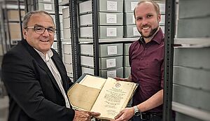 Hans-Bernd Köppen und Lennart Metken präsentieren einen Ordner mit alten Dokumenten. Neben ihnen befinden sich Metallregale gefüllt mit Dokumentenschachteln. Hans-Bernd Köppen und Lennart Metken präsentieren einen Ordner mit alten Dokumenten. Neben ihnen befinden sich Metallregale gefüllt mit Dokumentenschachteln.