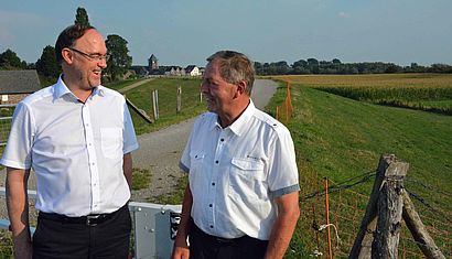 Weihbischof Rolf Lohmann und Deichgräf Herbert Scheers stehen auf einem Deich, der mit Gras bedeckt ist und reden miteinander. Im Hintergrund ist der Kirchturm von Dornick zu sehen.