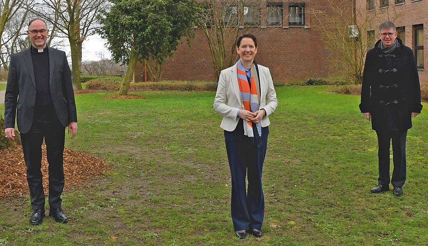 Eine Frau und zwei Männer stehen auf einer Wiese vor einem Gebäude.