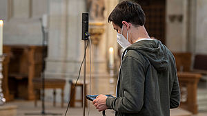 Ein junger Mann hält einen Tablet-Computer. Im Hintergrund ist eine Mevo-Kamera im Kirchenraum zu sehen.
