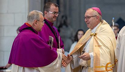 Ein Bischof reicht einem von zwei Domkapitularen die Hand.