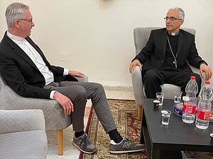 Zwei Männer in klerikaler Kleidung sitzen sich in hellgrauen Sesseln gegenüber und führen ein Gespräch. Beide tragen dunkle Anzüge mit weißem Kollarhemd; einer hat graue Hose und Sportschuhe, der andere einen langen silbernen Kreuzanhänger. Zwischen ihnen steht ein niedriger schwarzer Tisch. Der Raum ist schlicht eingerichtet, mit hellem Boden und einem Teppich unter dem Tisch.