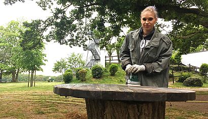 Carina Nitsch trägt Arbeitskleidung und schleift mit einer Maschine einen Tisch auf dem Außengelände des Michaelturms ab.