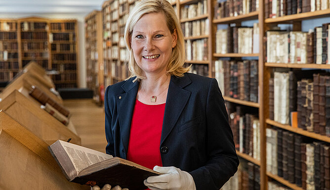 Eine Frau steht in einer alten Bibliothek und hält ein Buch in den Händen.
