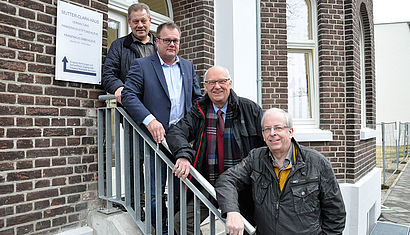 Vier Männer stehen auf einer Treppe, die nach links zum Eingang eines kernsanierten Hauses führt. Auf einem Schild links steht der Name Mutter-Clara-Haus.