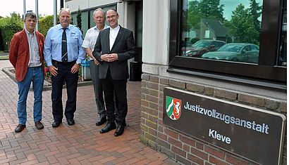 Vier Männer stehen vor einem Gebäude, an dem ein Schild mit der Aufschrift "Justizvollzugsanstalt Kleve" zu sehen ist.